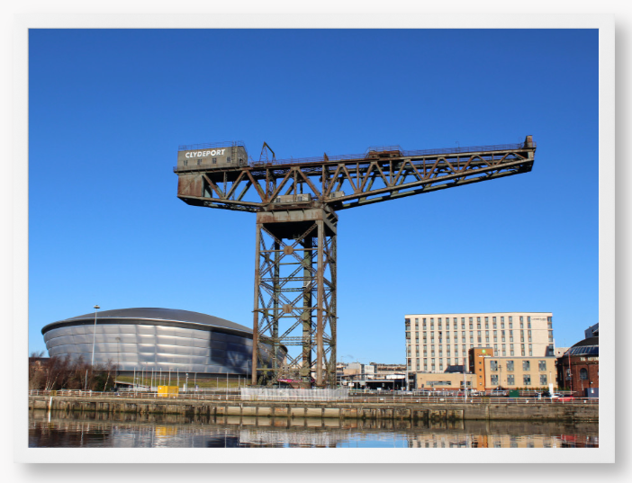 Framed Photo of a Industrial crane with modern buildings in the background under a clear blue sky.