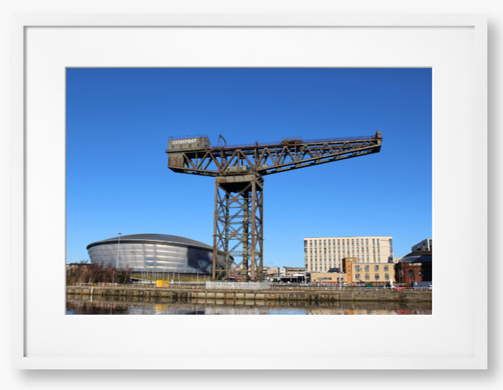 Framed Photo of a Industrial crane with modern buildings in the background under a clear blue sky.