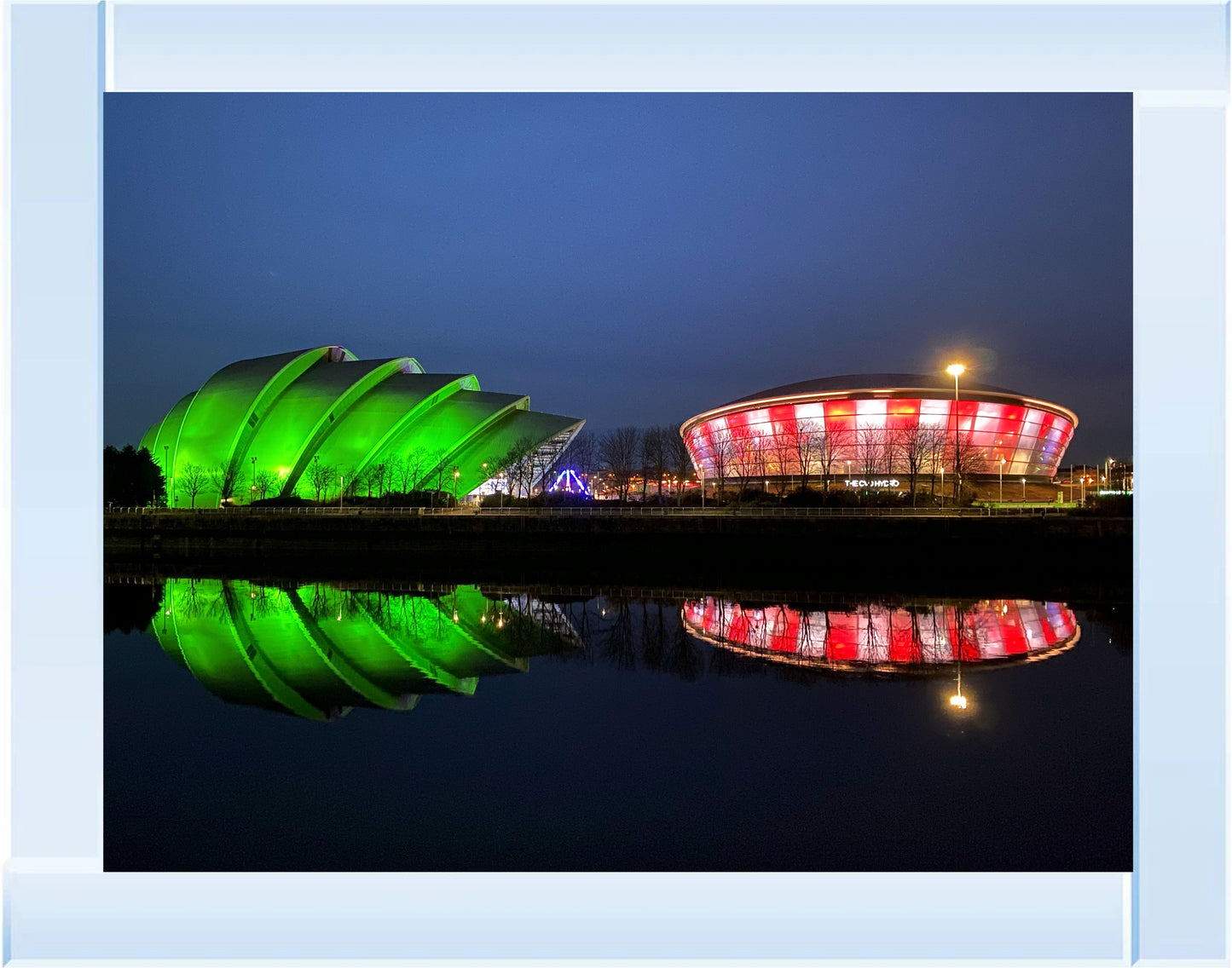 Two modern buildings, one green and one red and white, reflected in water at night.