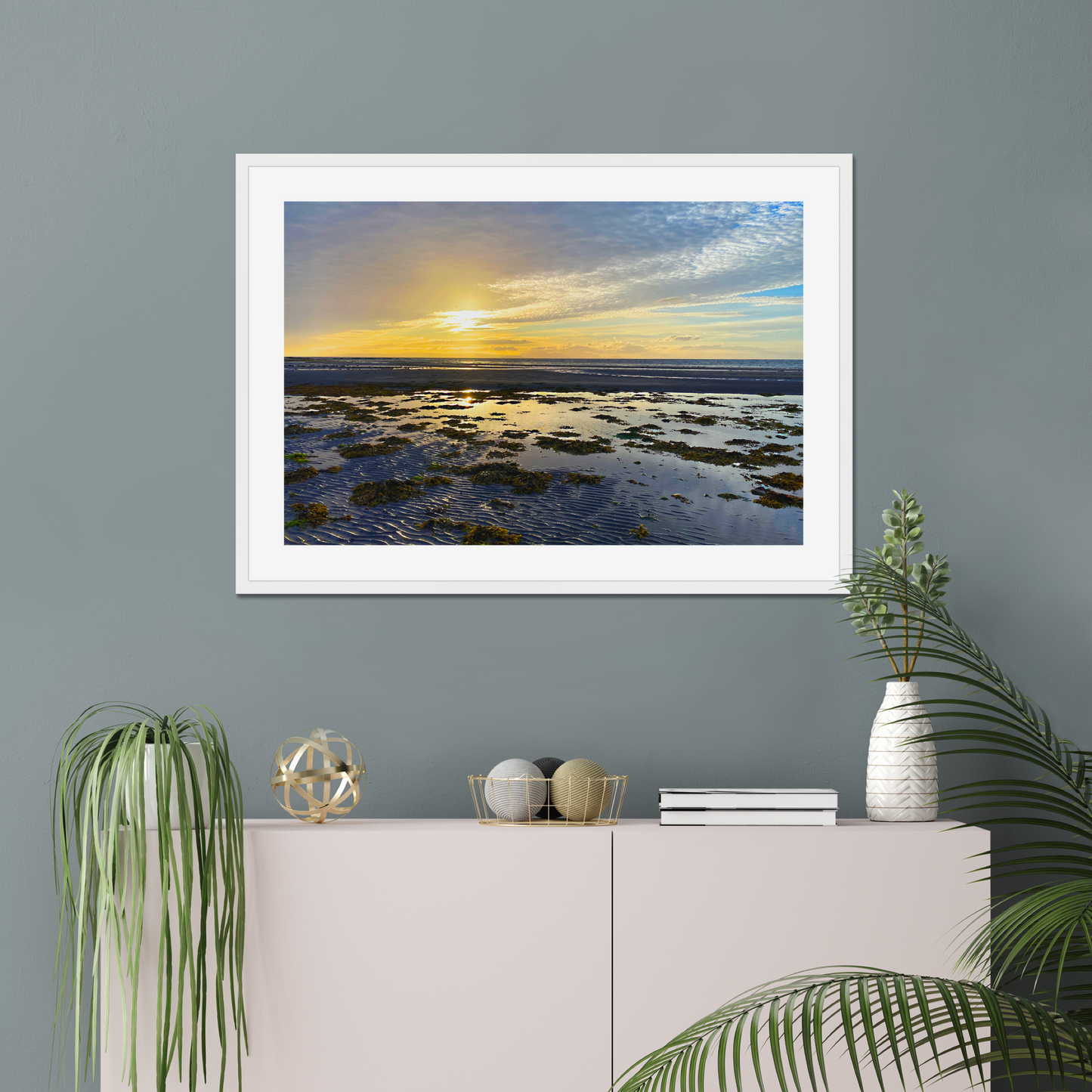 Framed beach landscape print on a gray wall above a white cabinet with decorative items.