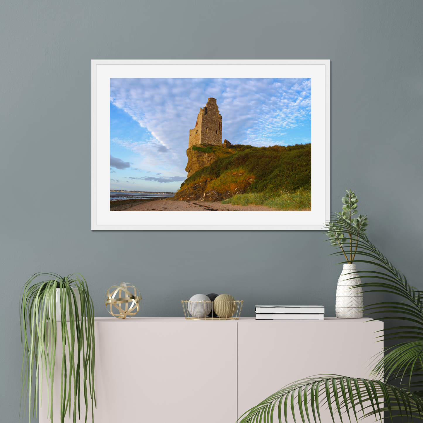 Framed picture of a coastal scene with a castle on a wall above a white cabinet with decorative items.