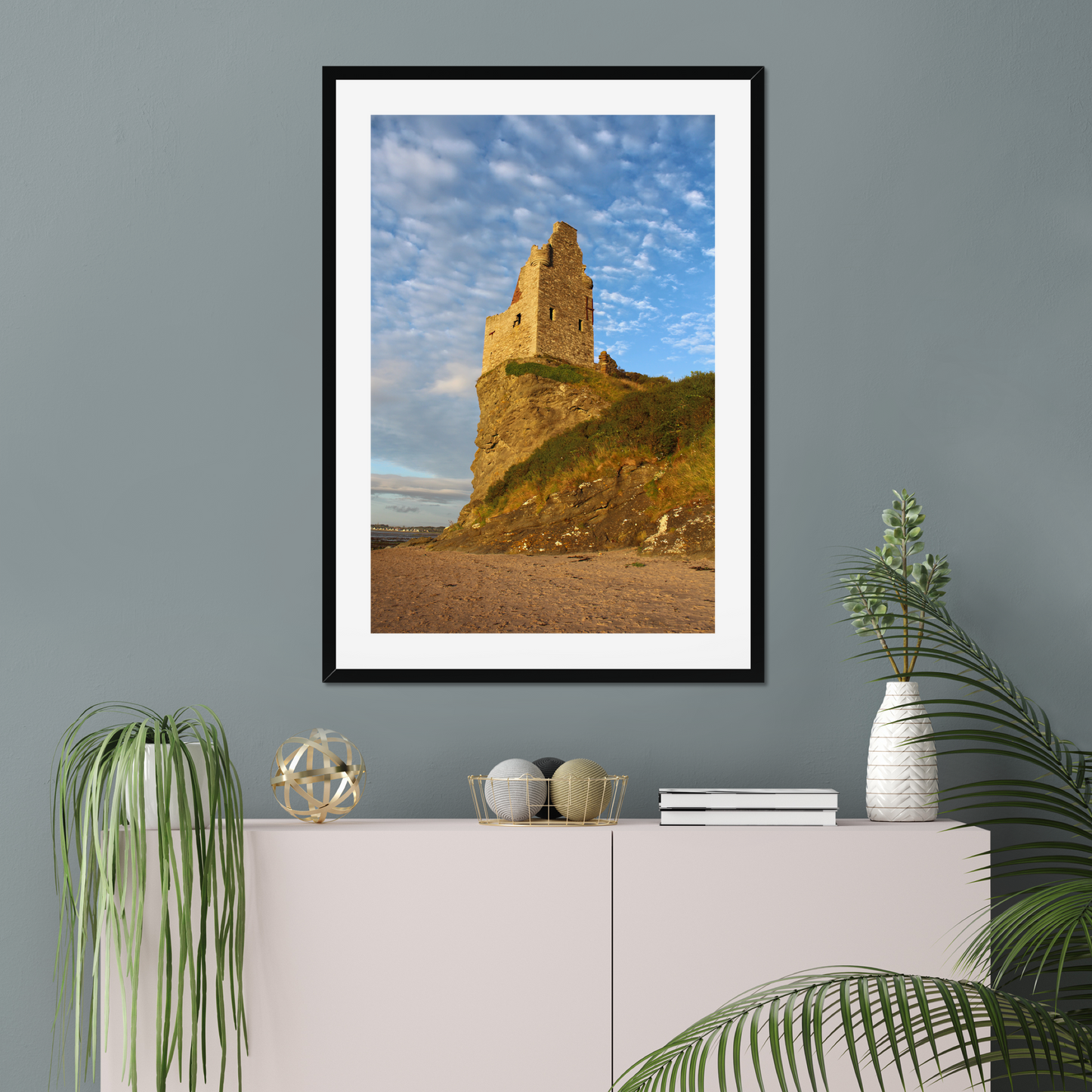 Framed photograph of a coastal scene with a castle on a gray wall above a white cabinet.