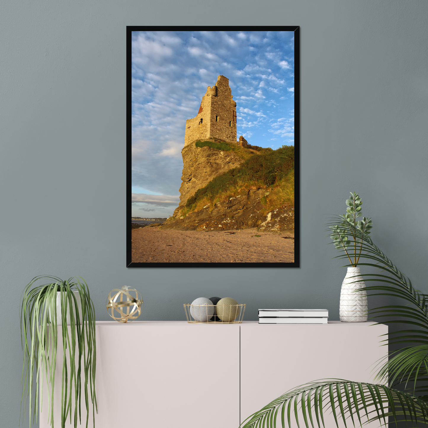 Framed artwork of a castle on a cliff against a blue sky, displayed on a wall above a white cabinet with decorative items.