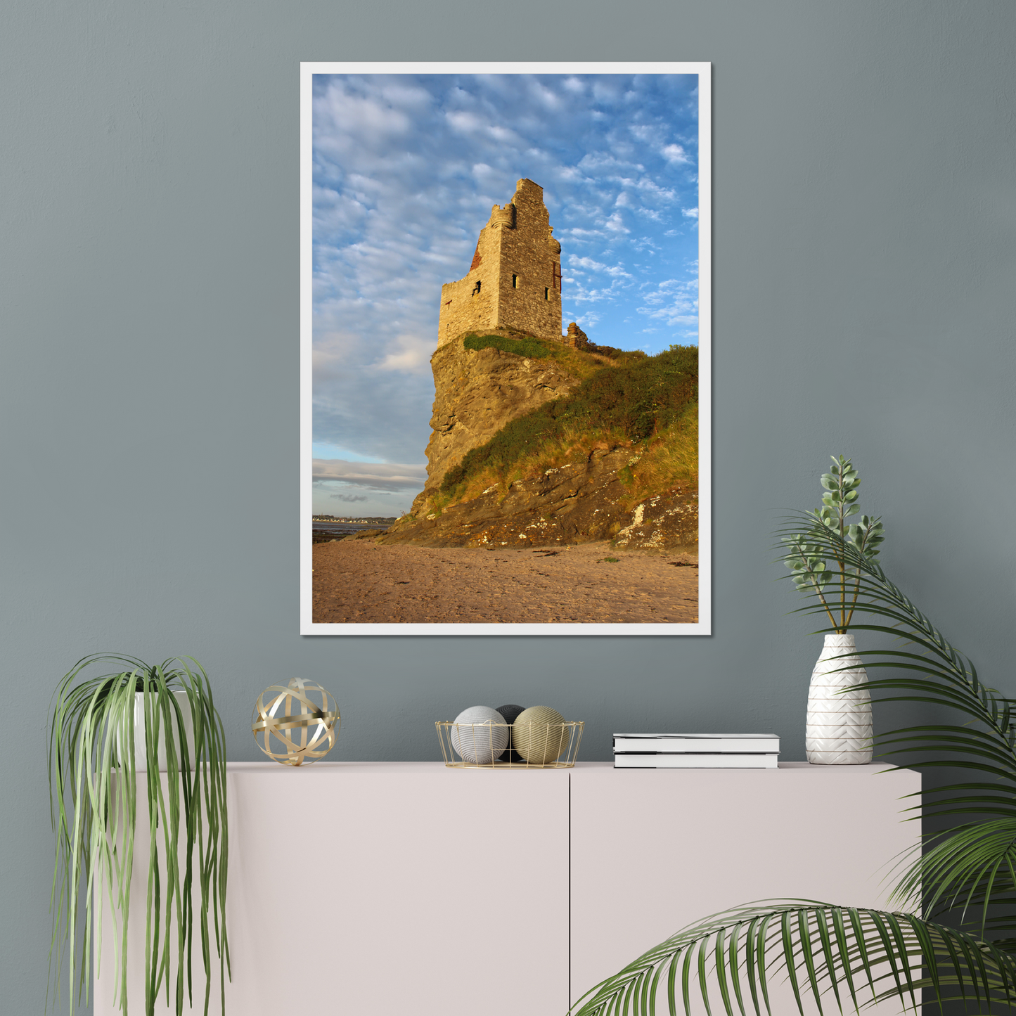 Framed photograph of a castle on a hill against a blue sky, displayed on a wall above a white cabinet with decorative items.