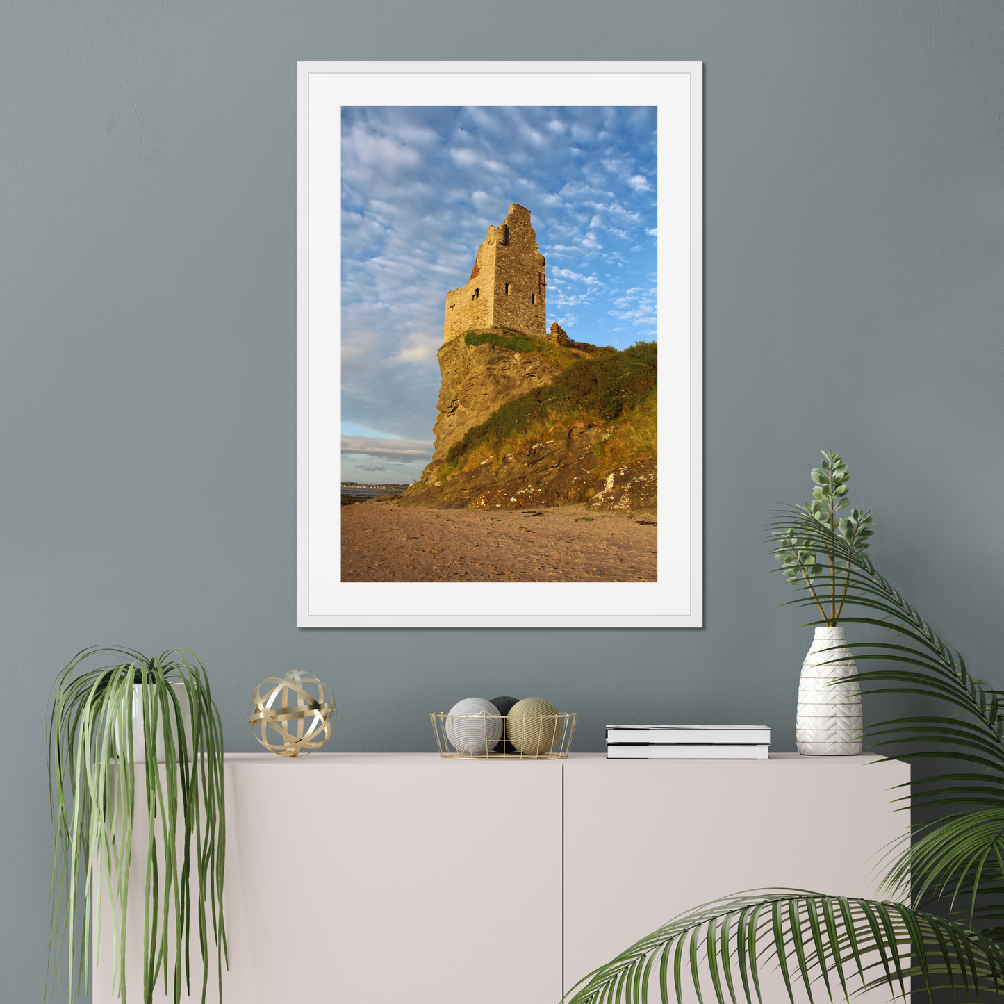 Framed photograph of a coastal scene with a castle on a gray wall above a white cabinet.