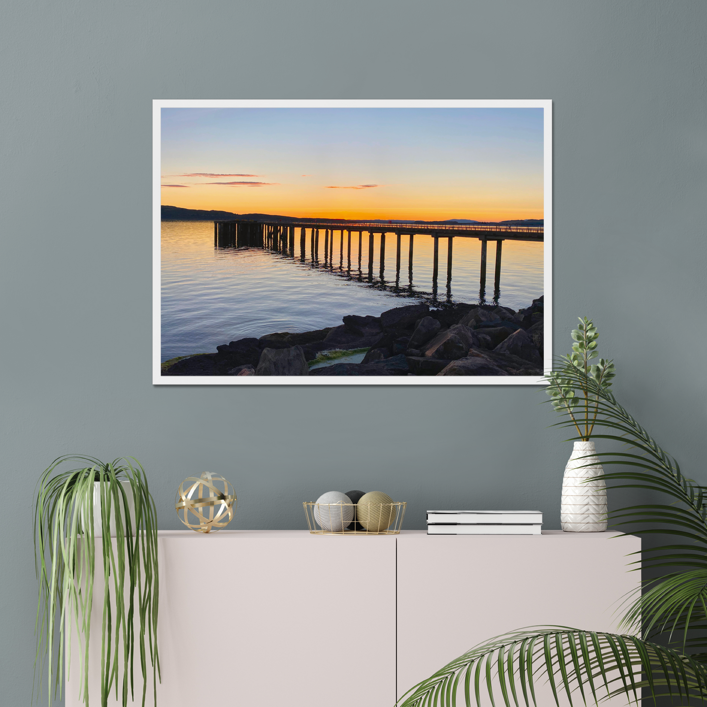 Framed artwork of a sunset over a pier on a gray wall above a white console table with decorative items.