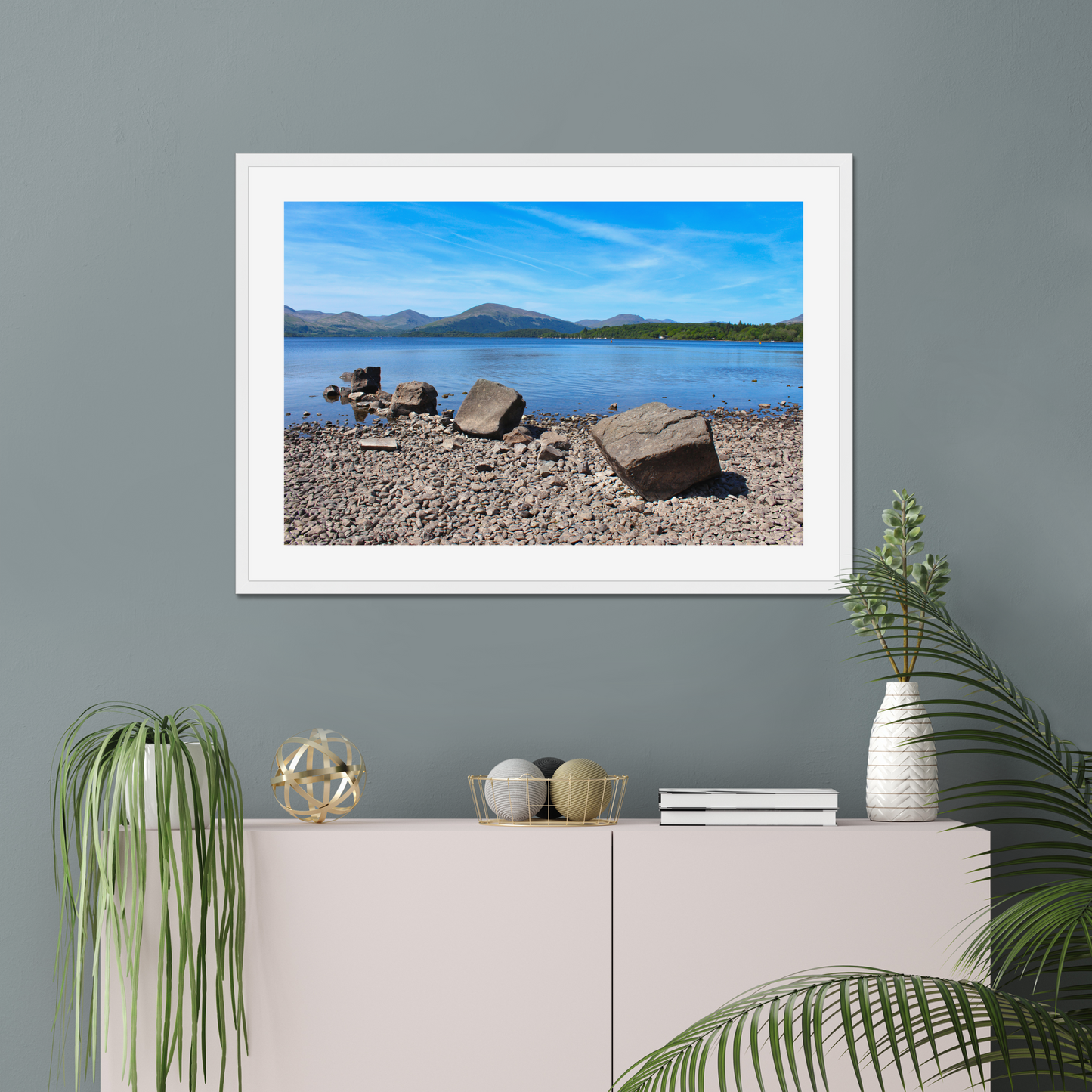 Framed landscape print of a lake and mountains on a gray wall above a white cabinet with decorative items.