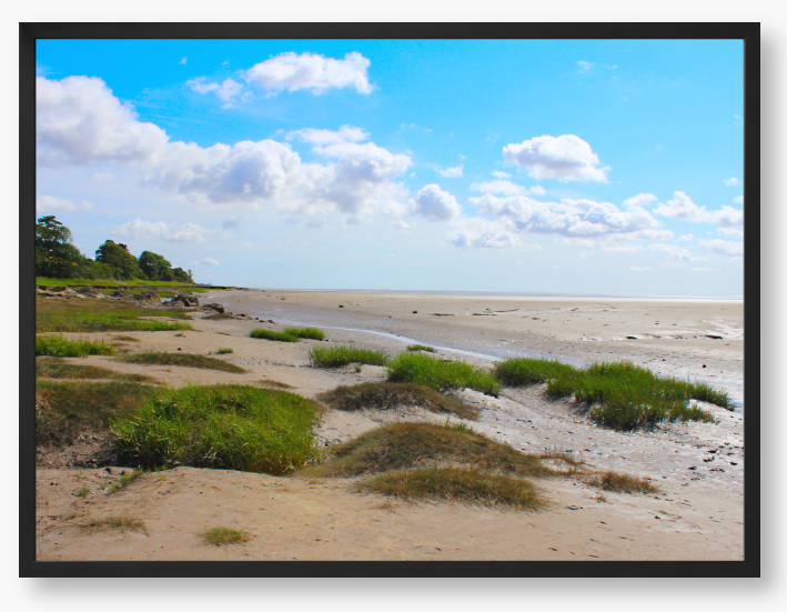 Framed photograph of a coastal landscape with sandy beach, grassy areas, and blue sky.