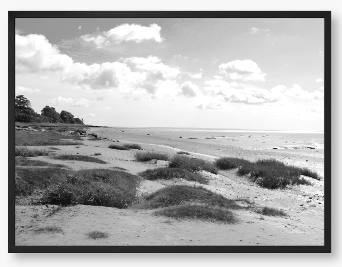 Black and White Framed photograph of a coastal landscape with sandy beach, grassy areas, and cloudy sky.