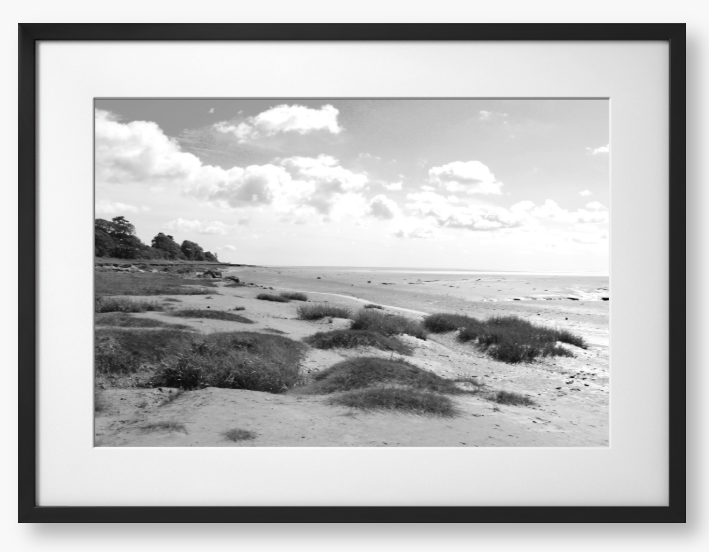 Black and White Framed photograph of a coastal landscape with sandy beach, grassy areas, and cloudy sky.