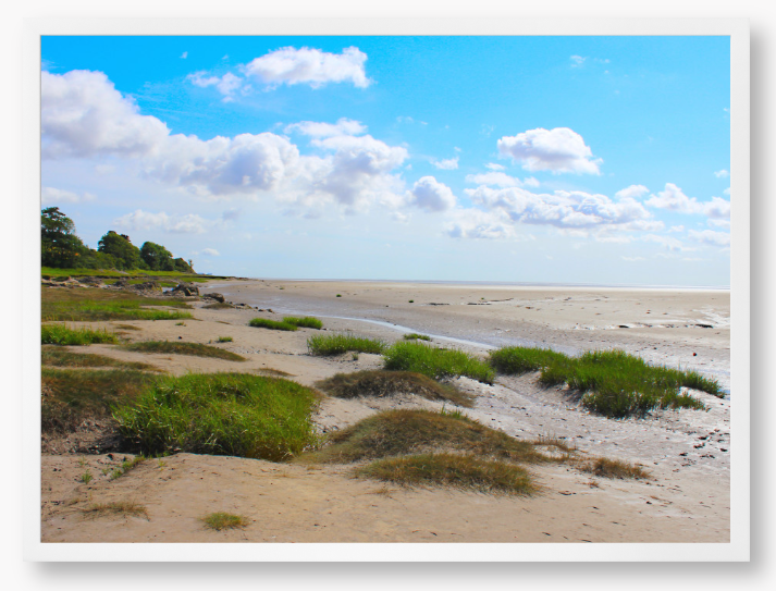 Framed photograph of a coastal landscape with sandy beach, grassy areas, and blue sky.