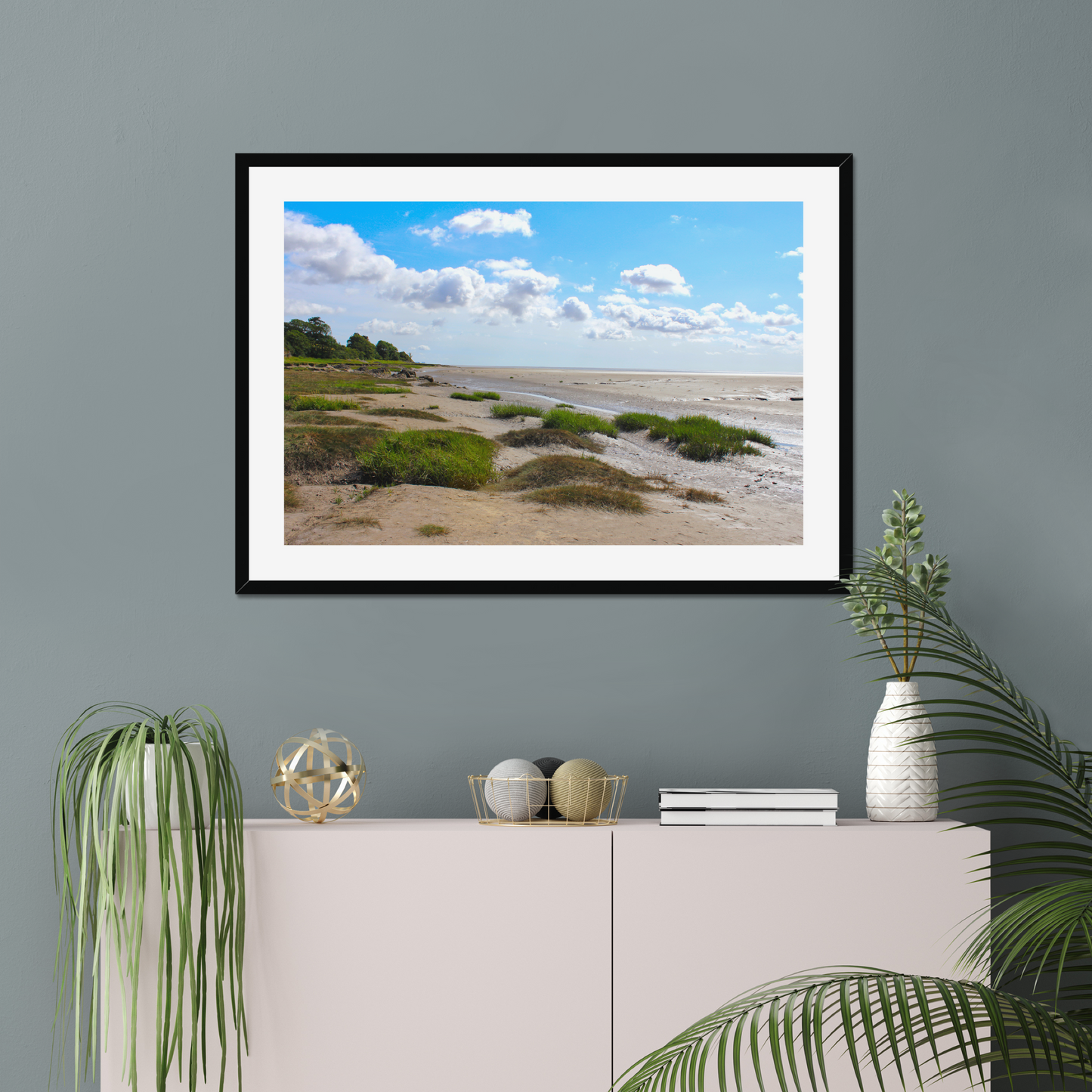 Framed landscape print of a beach scene on a gray wall above a white cabinet with decorative items.