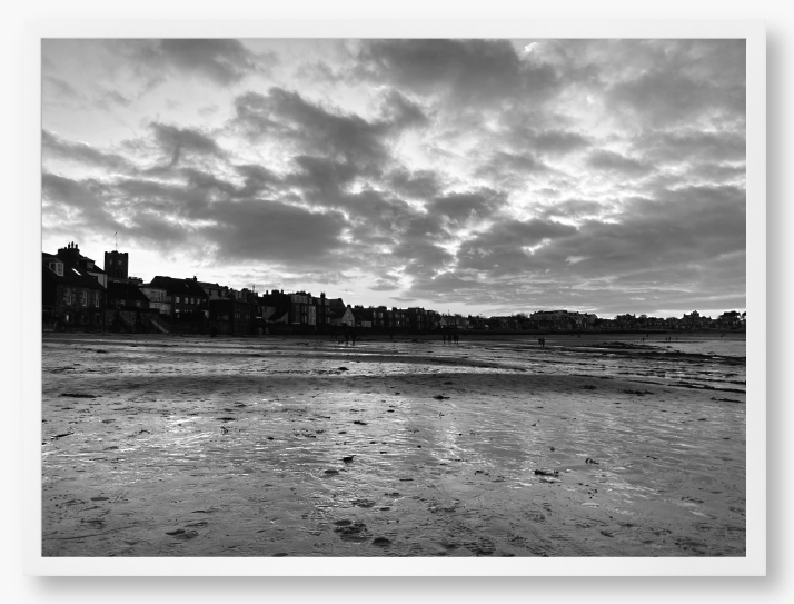 Black and white framed photograph of a coastal scene with buildings and clouds.