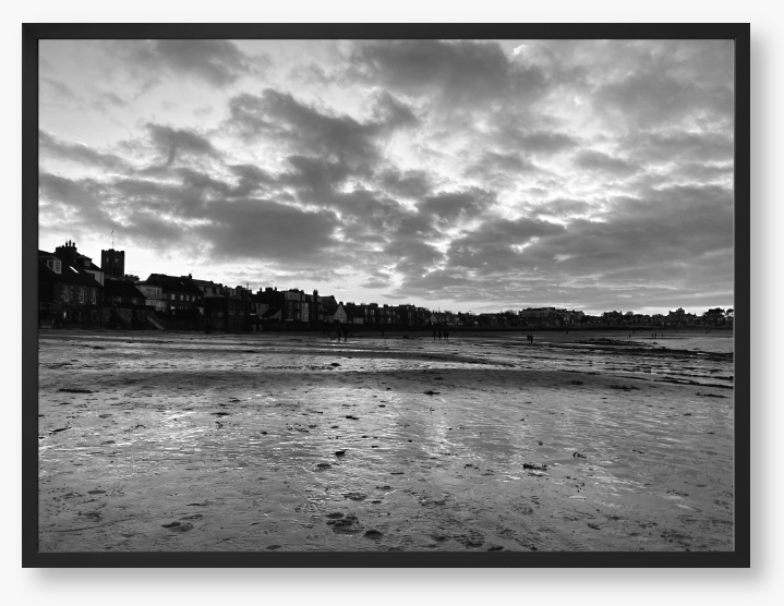 Black and white framed photograph of a coastal scene with buildings and clouds.