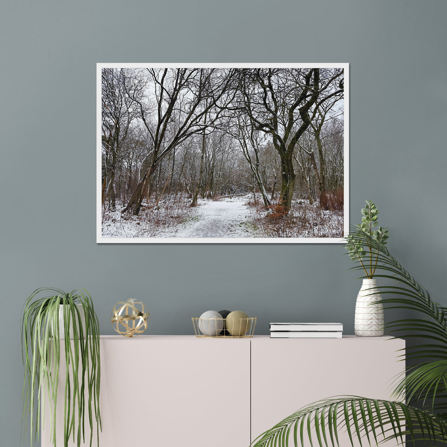 Framed photograph of a winter woodland scene with snow on the ground on a gray wall above a white cabinet with decorative items.