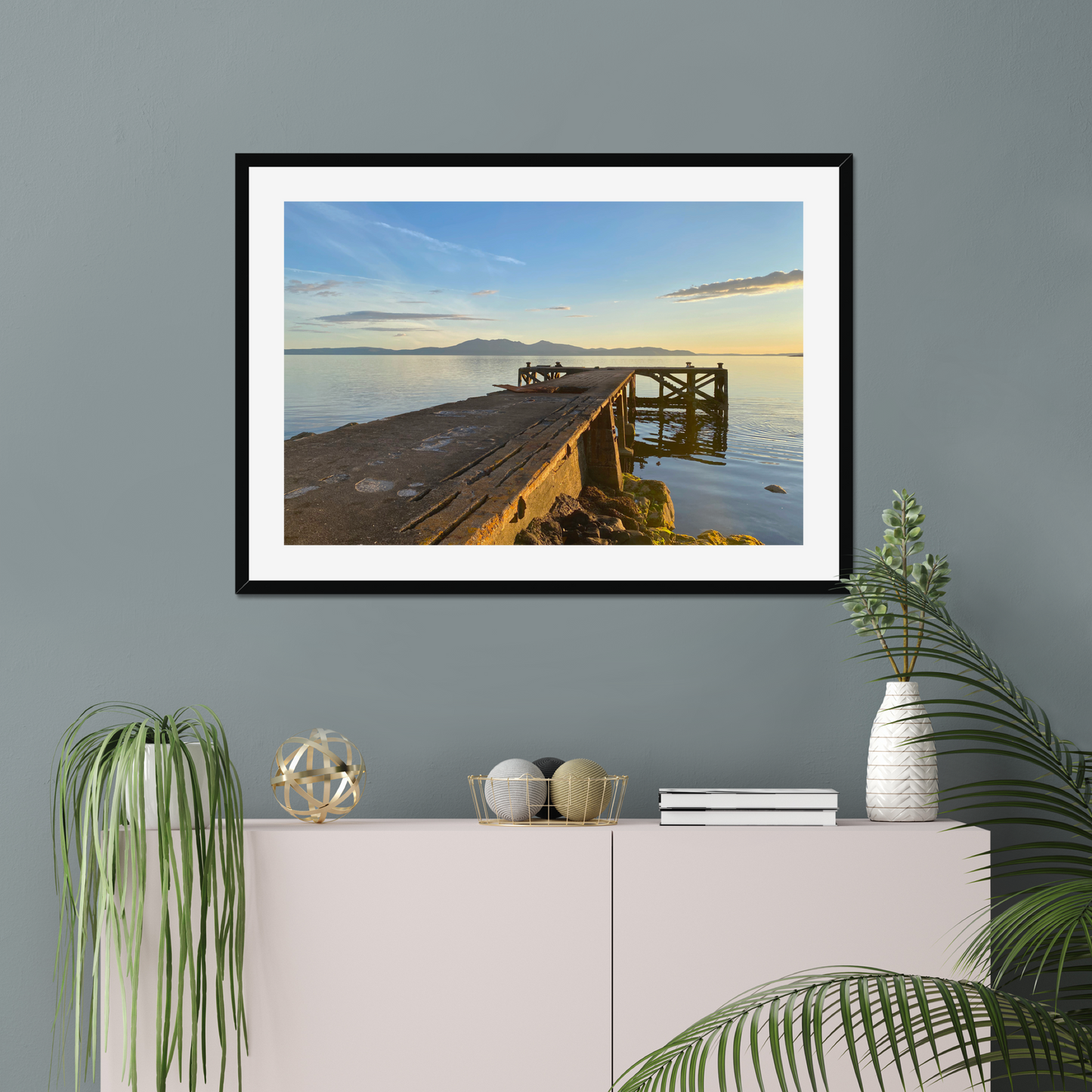 Framed artwork of a bridge at sunset on a gray wall above a white cabinet with decorative items.