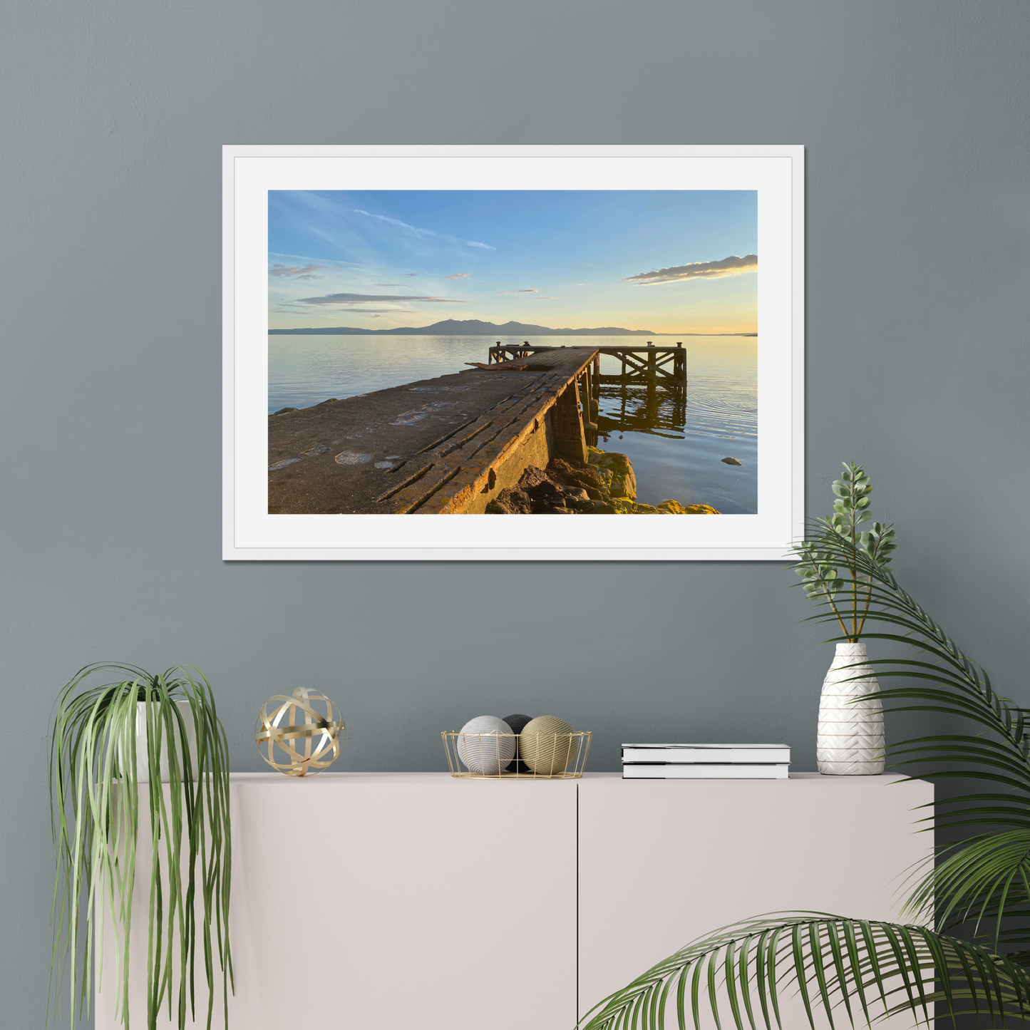 Framed photograph of a dock at sunset on a gray wall above a white cabinet with decorative items.