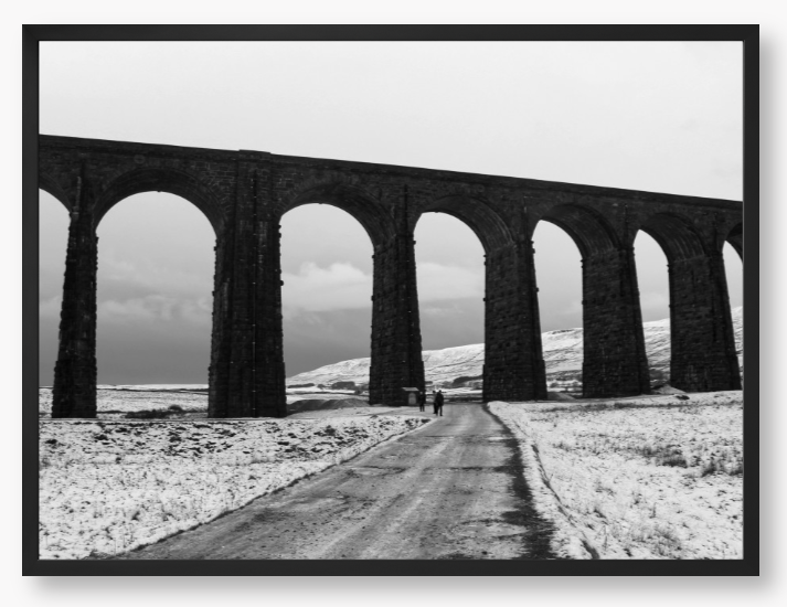 black and white Framed Photo of a Viaduct in a snowy landscape with people walking on a path below.