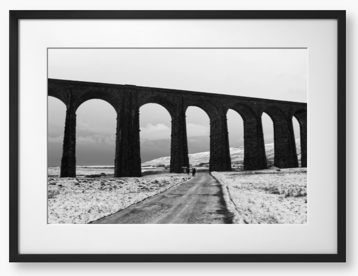 black and white Framed Photo of a Viaduct in a snowy landscape with people walking on a path below.