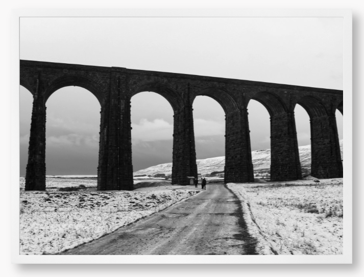 black and white Framed Photo of a Viaduct in a snowy landscape with people walking on a path below.