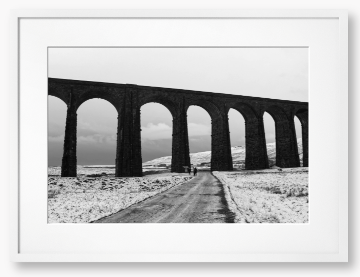 black and white Framed Photo of a Viaduct in a snowy landscape with people walking on a path below.