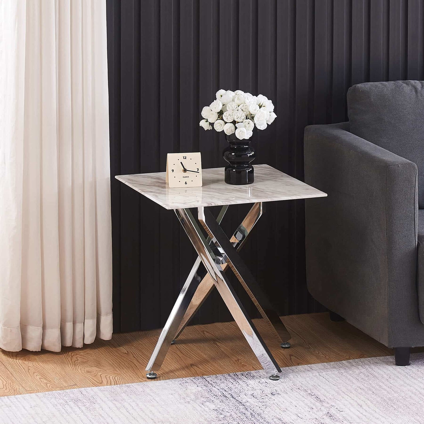 Small marble table with a clock and flower vase next to a gray sofa in a room with black curtains.