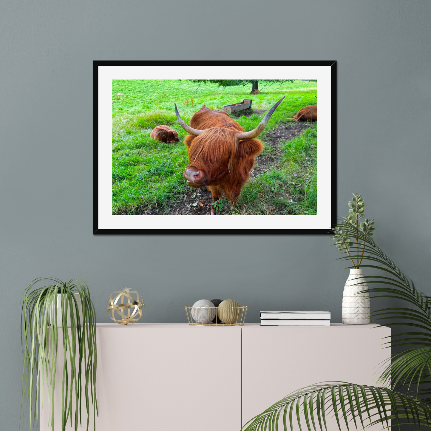Framed picture of a highland cow on a wall above a cabinet with decorative items.