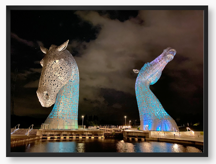 Framed photo of the Kelpies horse sculptures in Falkirk, Scotland, illuminated at night with a colourful light display.