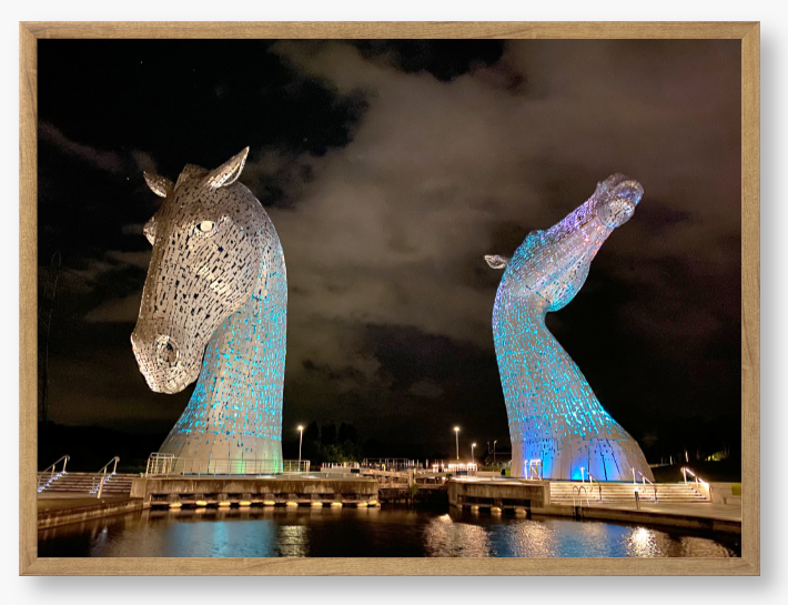 Framed photo of the Kelpies horse sculptures in Falkirk, Scotland, illuminated at night with a colourful light display.