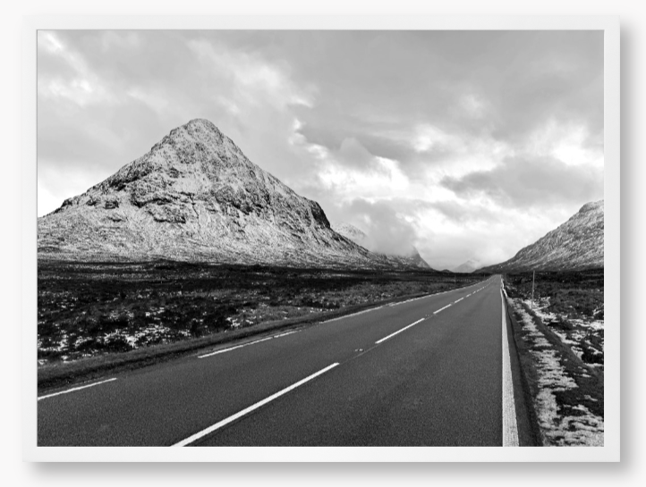Black and White Framed Photo of a road leading towards snow-capped mountains under a cloudy sky