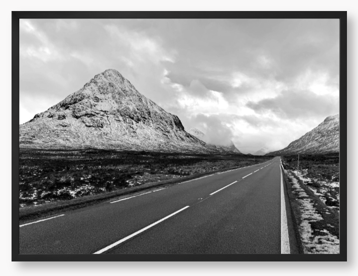 Black and White Framed Photo of a road leading towards snow-capped mountains under a cloudy sky