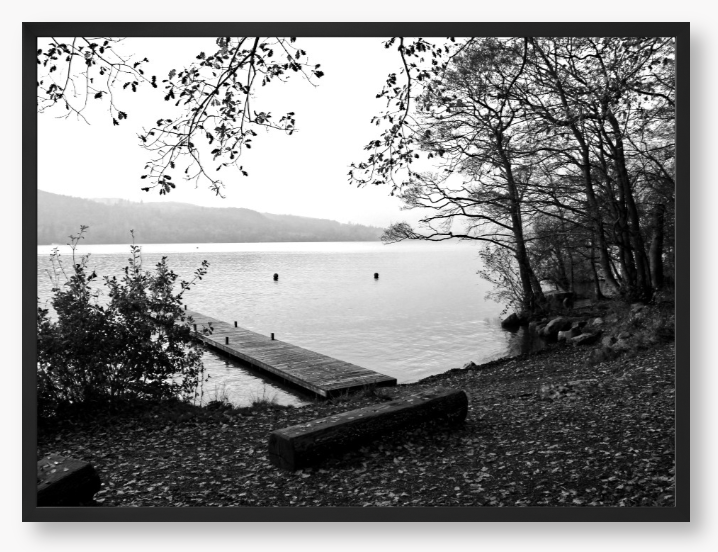 Black and white framed photo of a scene of a lake with a wooden jetty and trees.