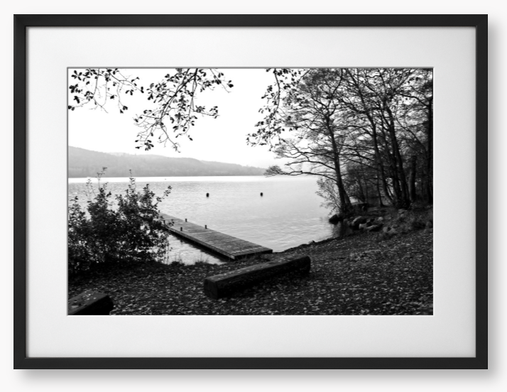 Black and white framed photo of a scene of a lake with a wooden jetty and trees.
