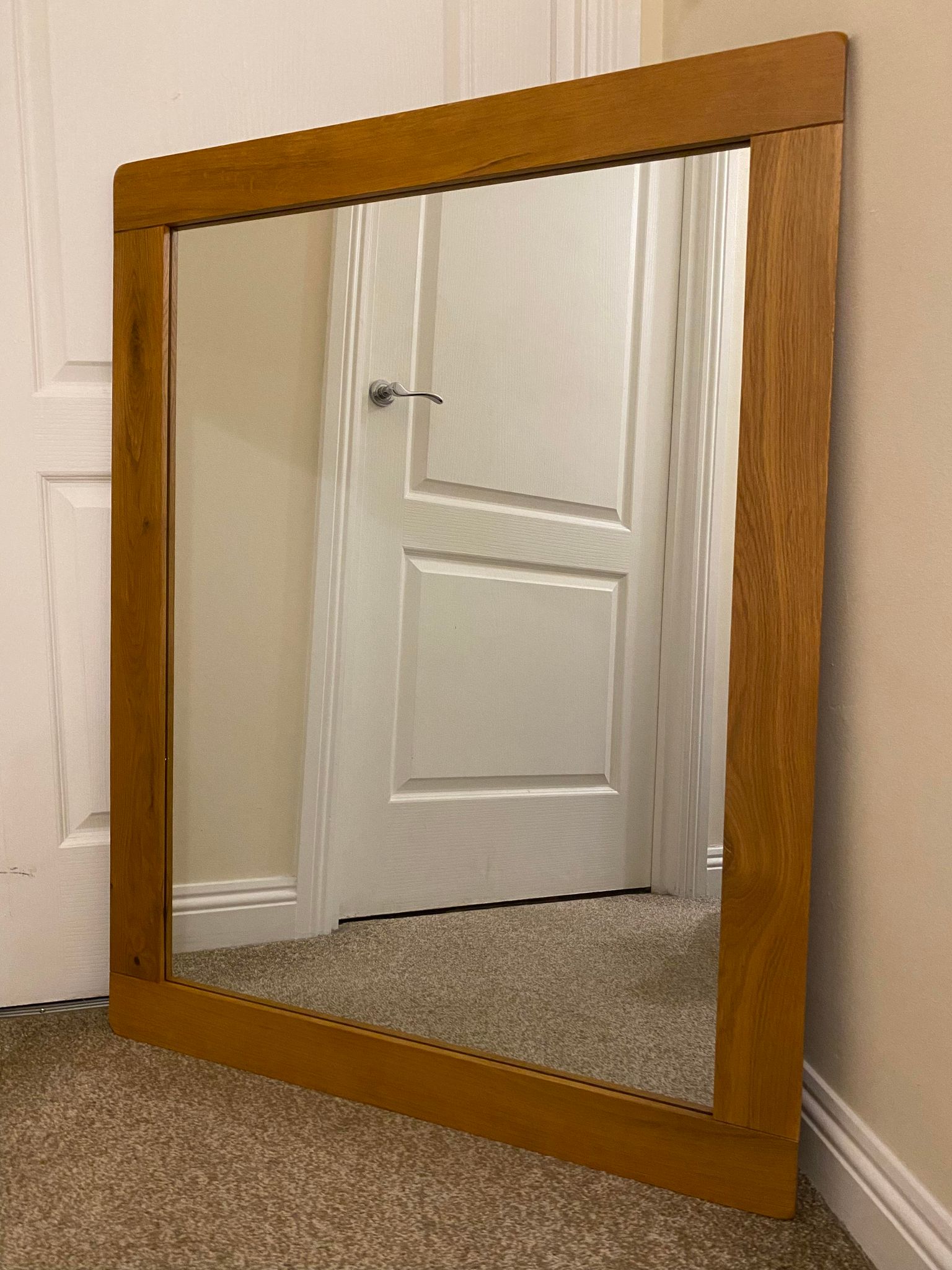 A large rectangular oak mirror leaning against a wall, reflecting a door and part of a room.
