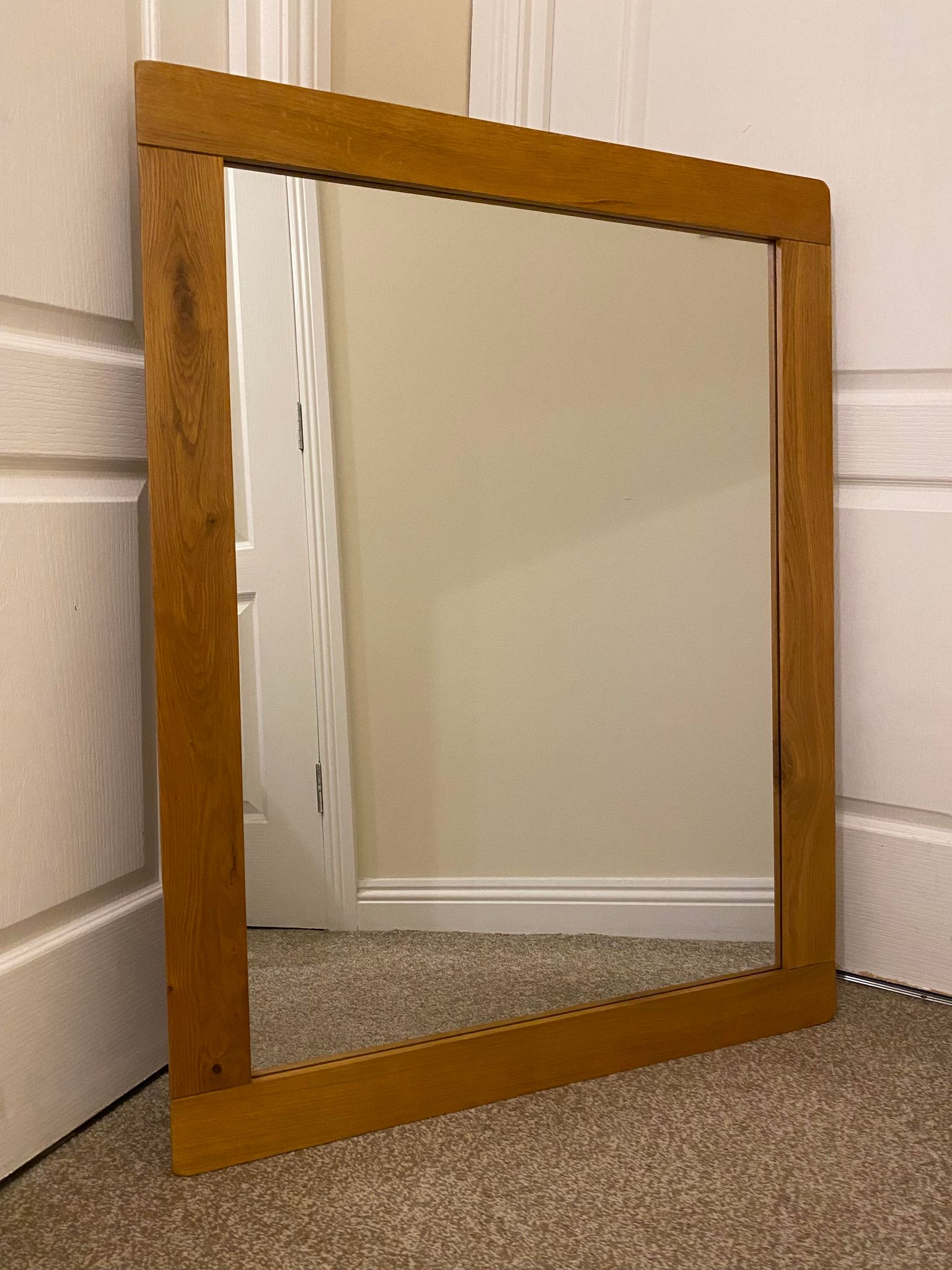 A large rectangular oak mirror leaning against a wall, reflecting a door and part of a room.