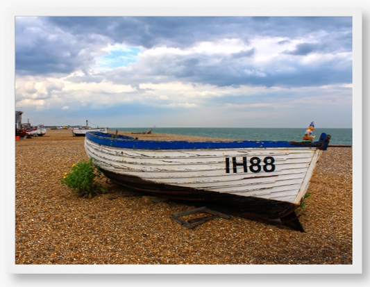 Framed Photo Of A White boat with blue trim on a pebbly beach under a cloudy sky