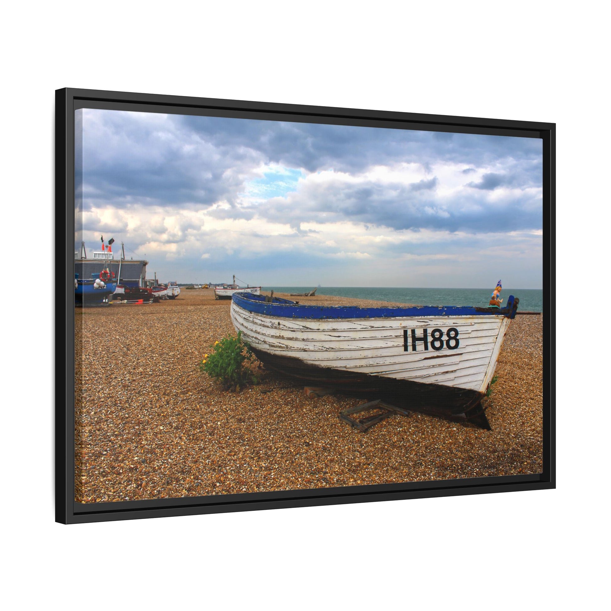 Framed photograph of a boat on a pebbly beach with a cloudy sky.