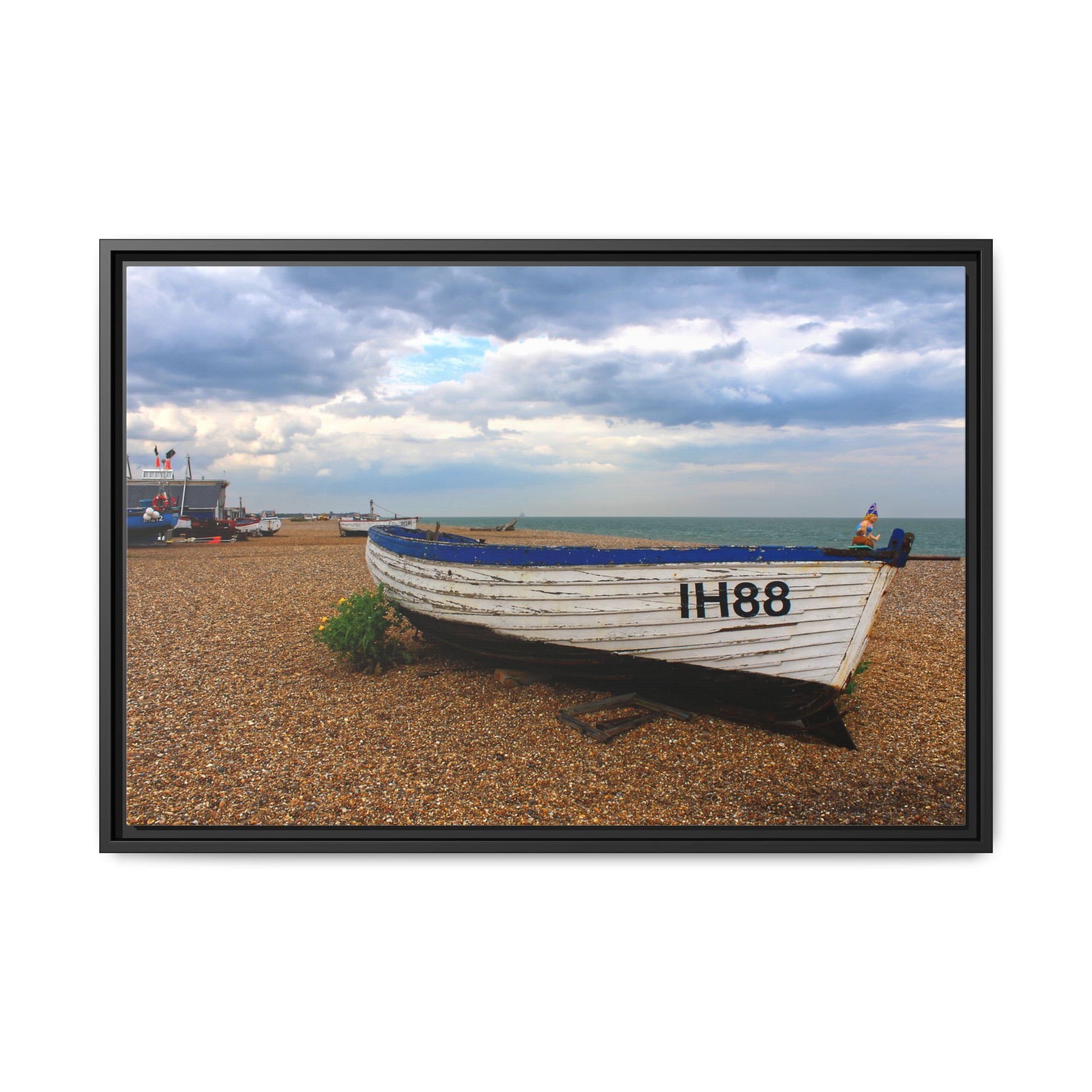 Framed photograph of a boat on a pebbly beach with a cloudy sky.