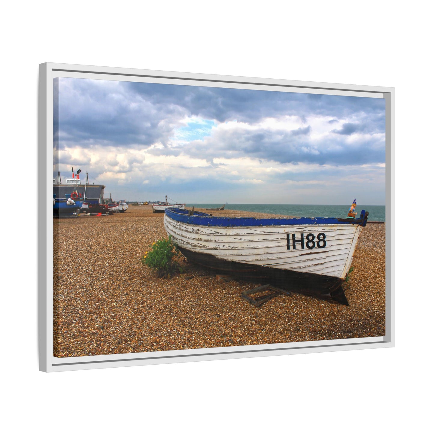 Framed photograph of a boat on a pebbly beach with a cloudy sky.