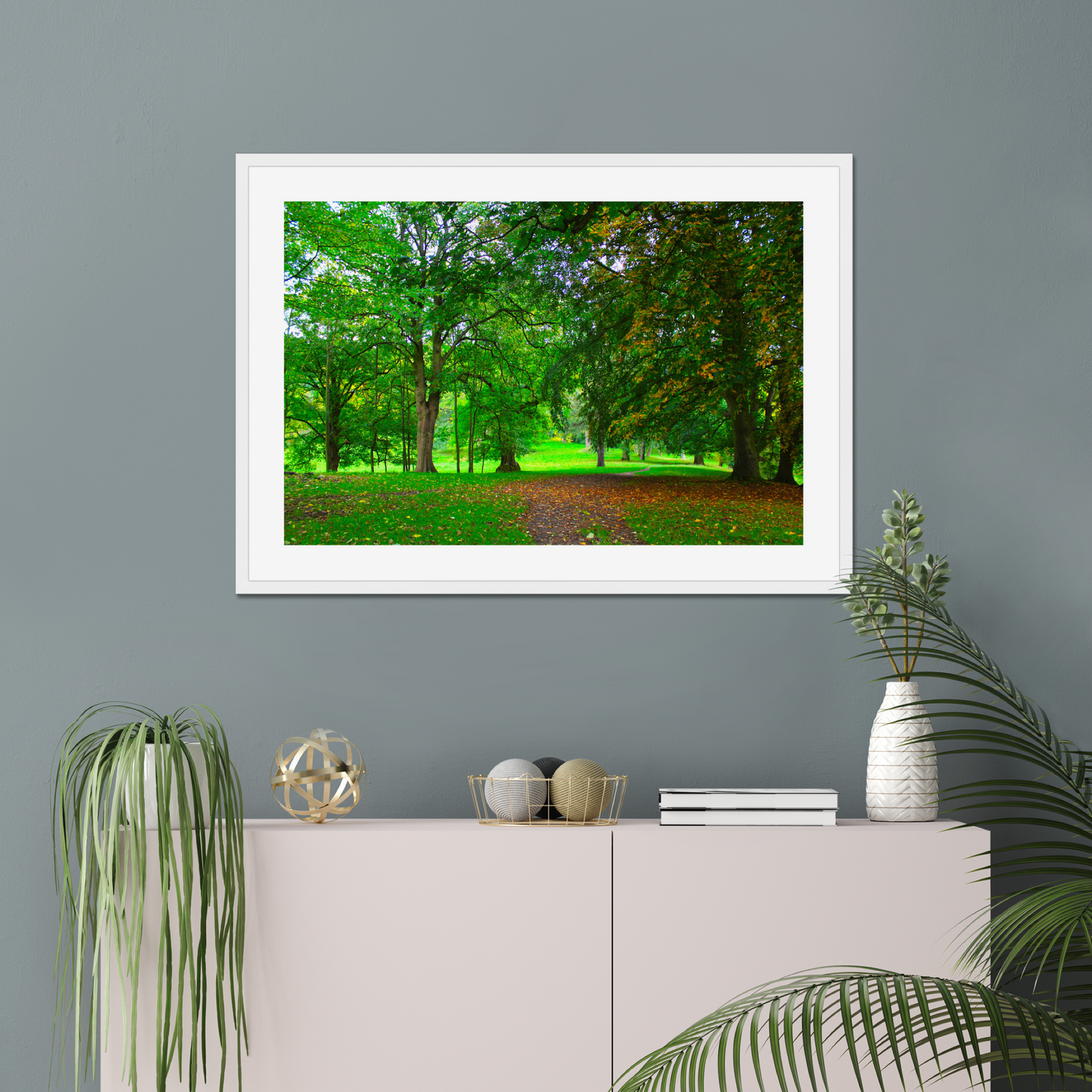 Framed nature print of a green forest scene on a gray wall above a white cabinet with decorative items.