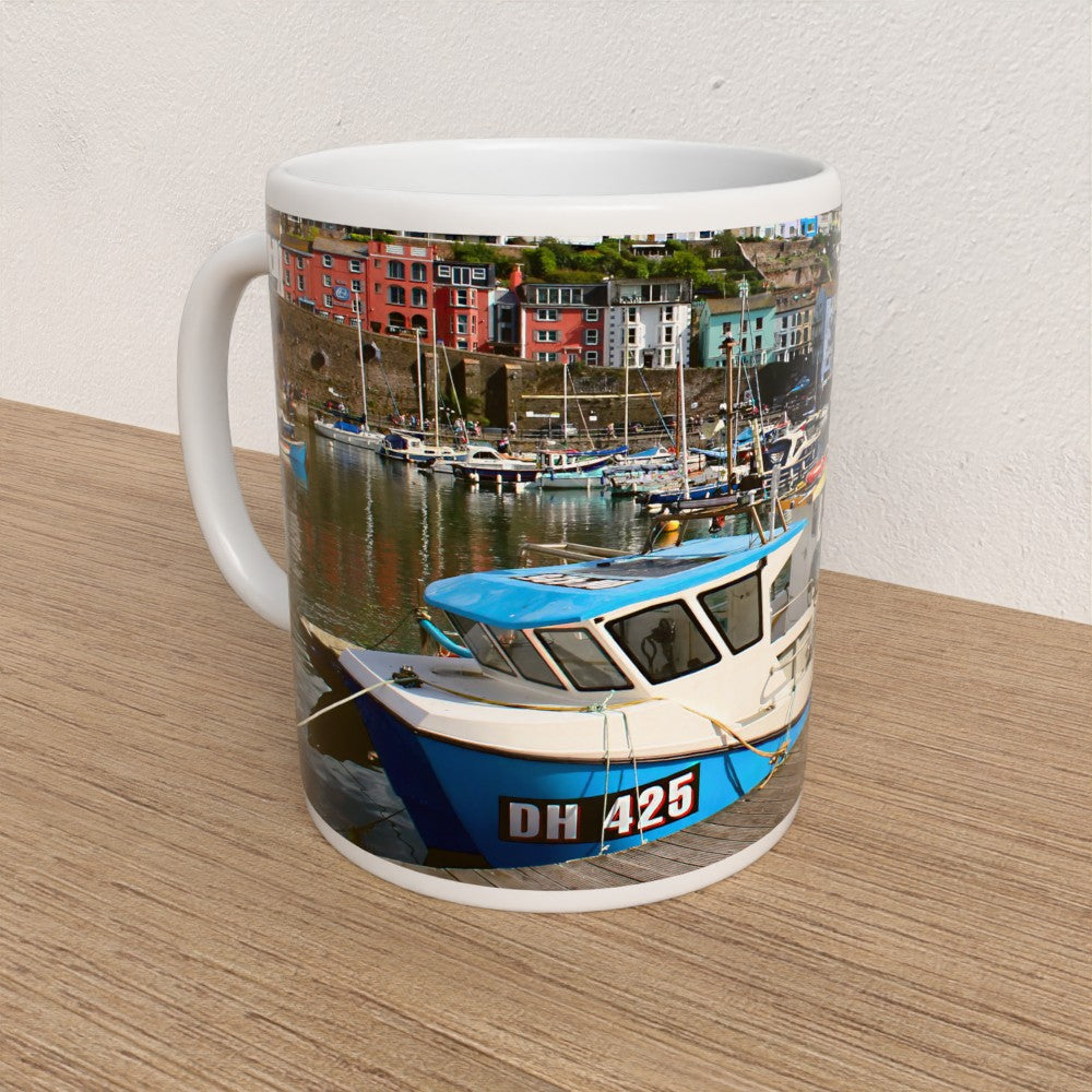 Mug with a boat and harbor scene printed on it, placed on a wooden surface.