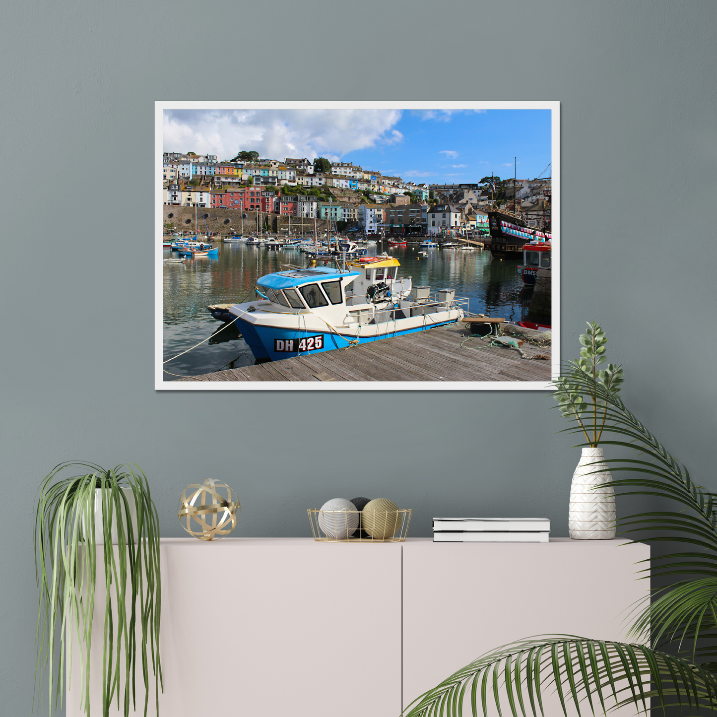 Framed photograph of a harbour scene on a wall above a cabinet with decorative items.