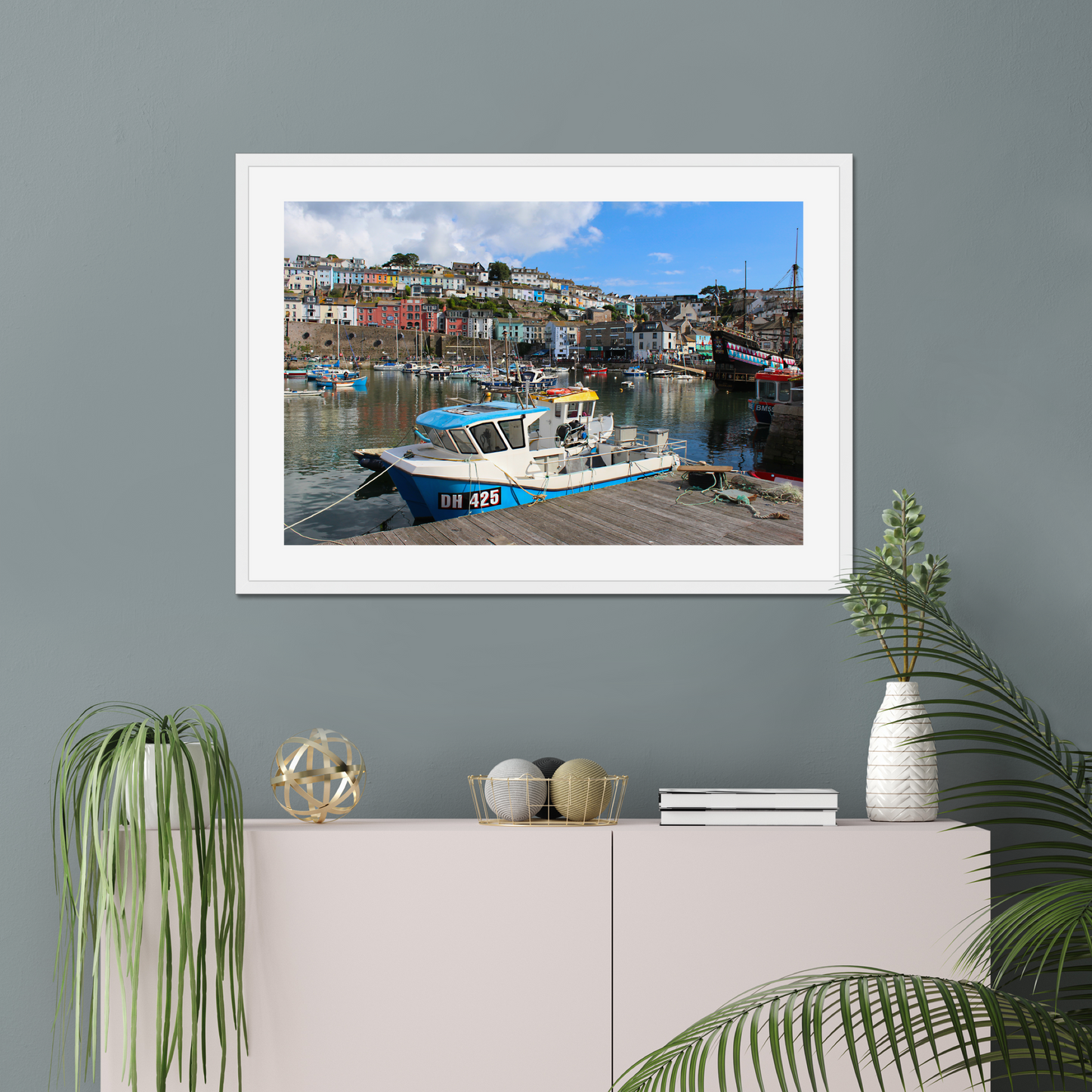 Framed photograph of a harbour scene on a wall above a cabinet with decorative items.