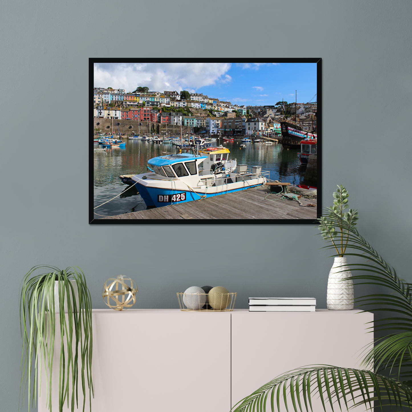 Framed photograph of a harbour scene on a wall above a cabinet with decorative items.