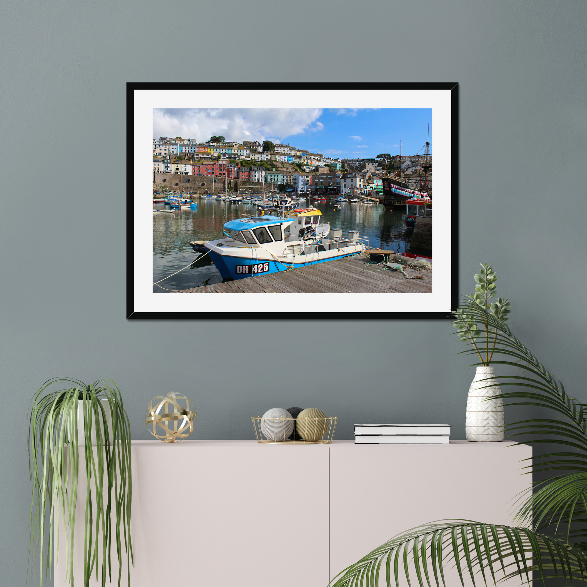 Framed photograph of a harbour scene on a wall above a cabinet with decorative items.