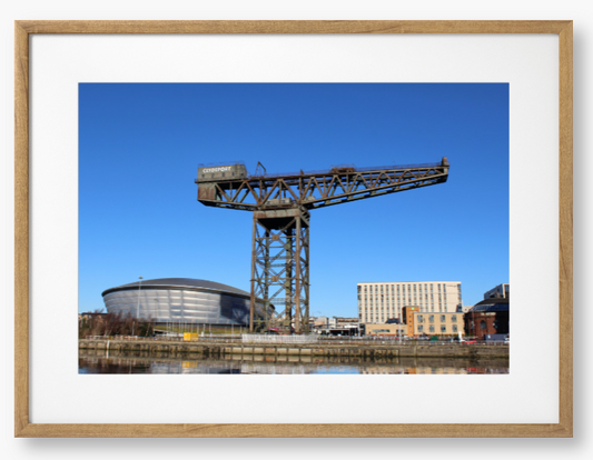 Framed Photo of a Industrial crane with modern buildings in the background under a clear blue sky.