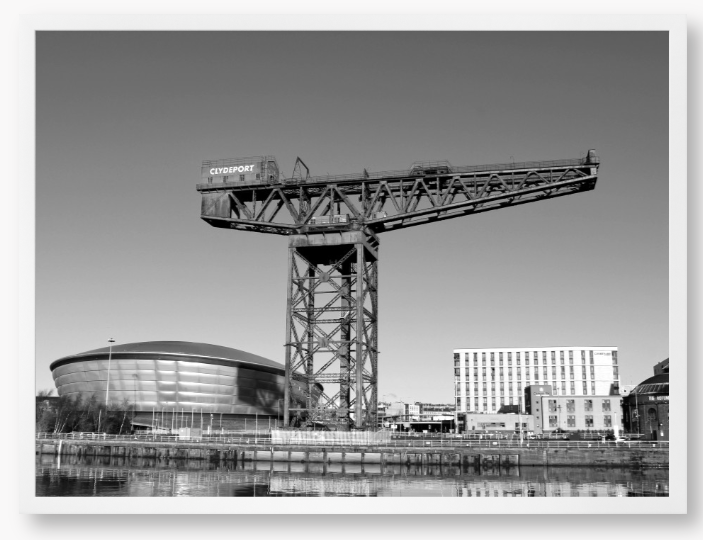 Black and white framed photo of a large crane with modern buildings in the background.