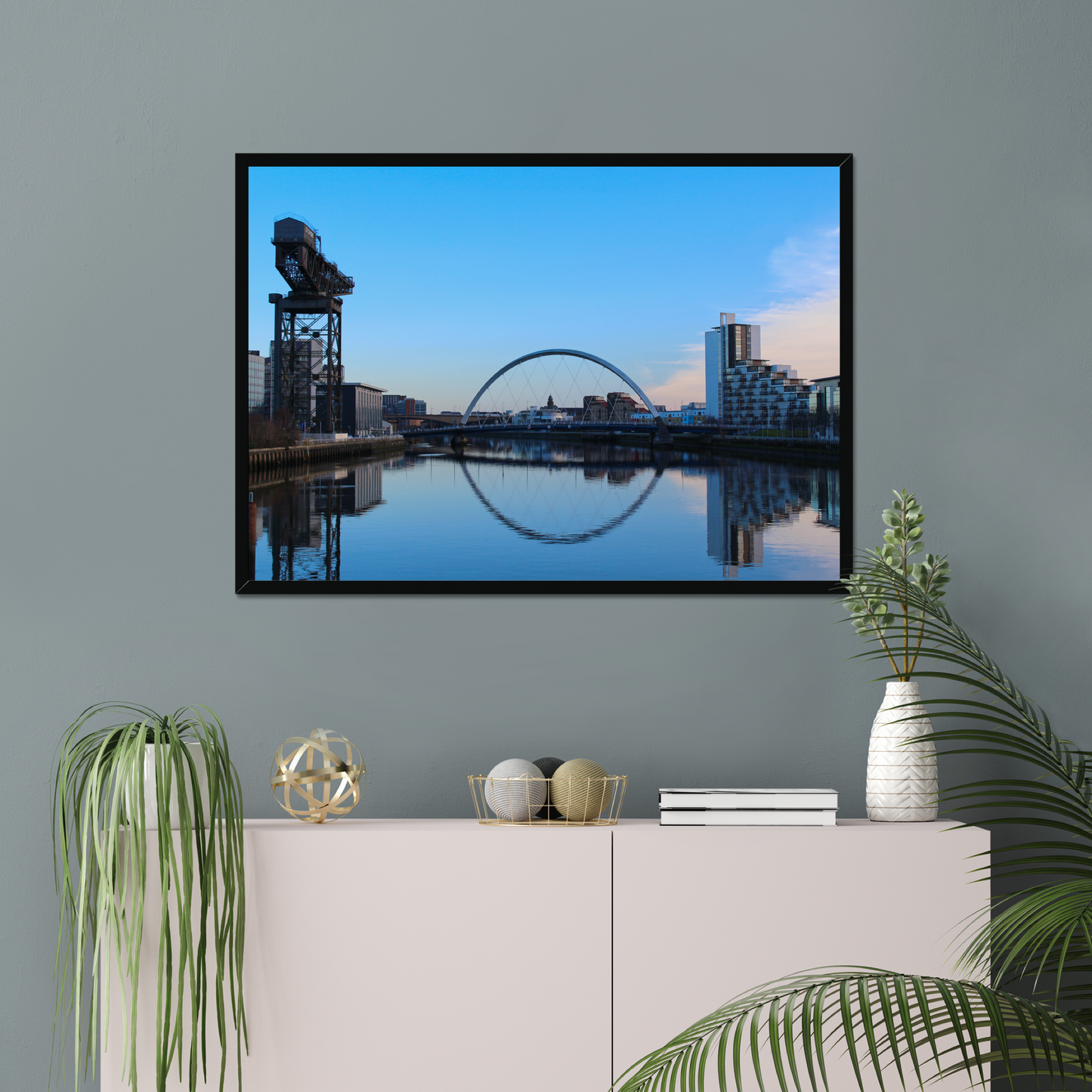 Framed cityscape with a bridge and skyline on a wall above a white cabinet with decorative items.