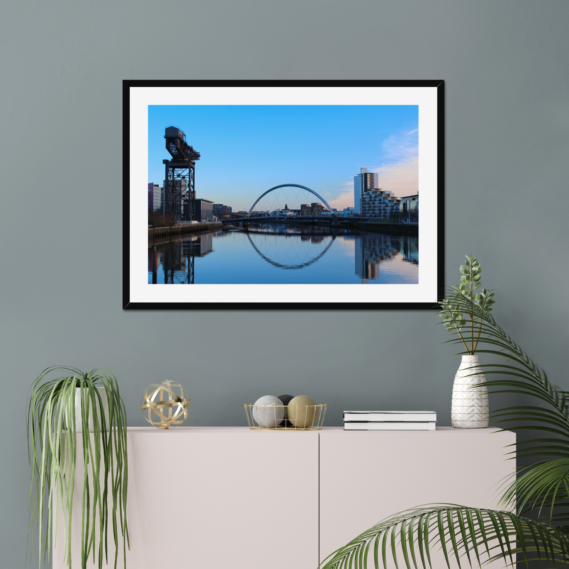 Framed artwork of a cityscape with a blue sky and reflection on water, displayed on a wall above a white cabinet with decorative items.