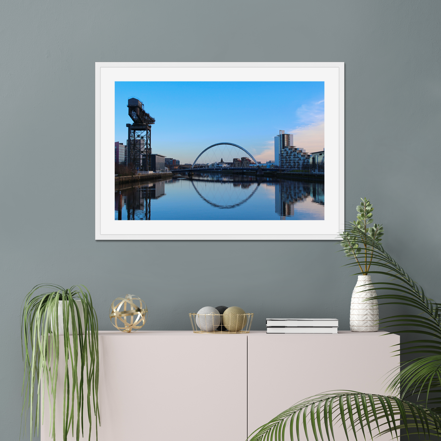 Framed picture of a cityscape with a river and skyline on a gray wall above a white cabinet with decorative items.